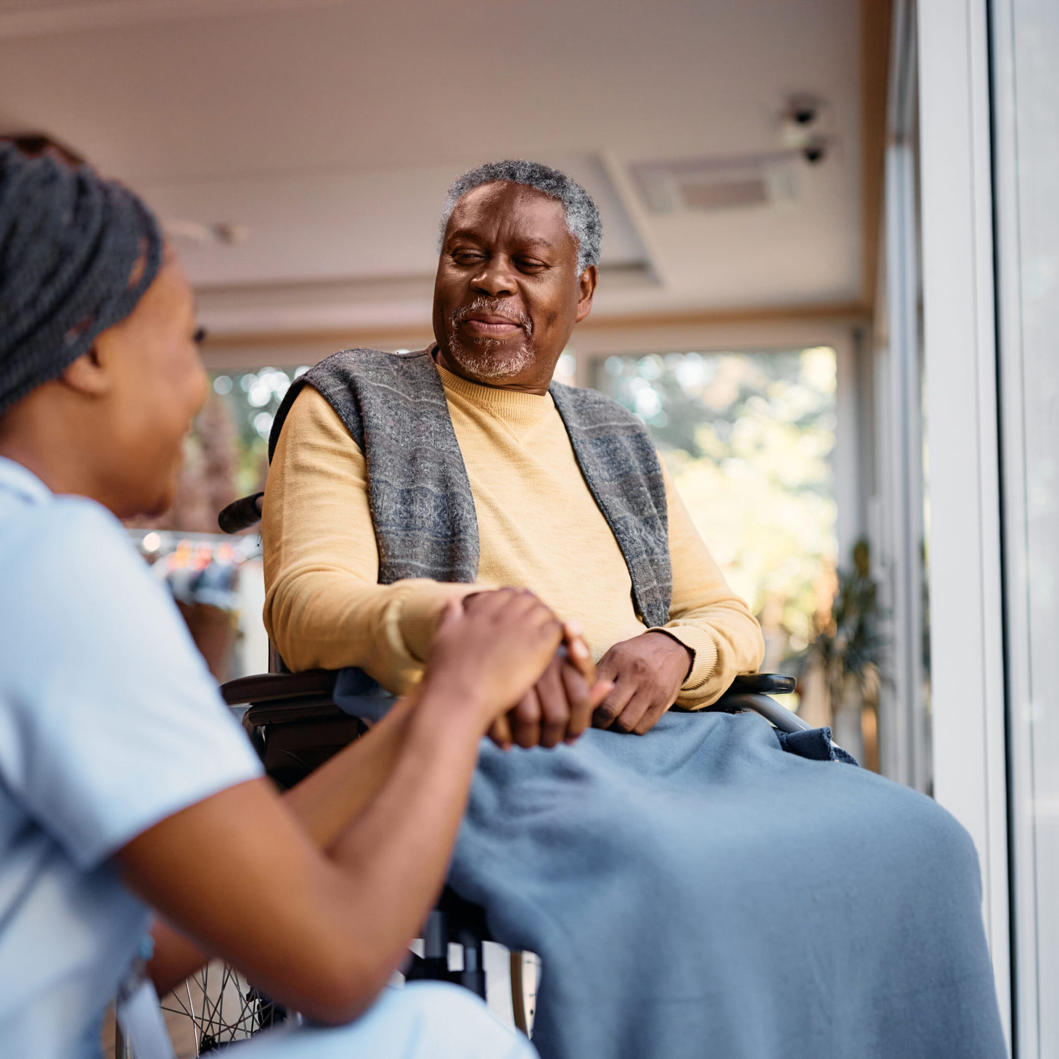 Caring African American nurse communicating with senior man in wheelchair while holding his hand in nursing home. Copy space.